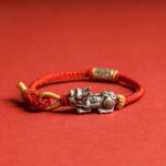 Sterling silver Pixiu red string bracelet side view with braided cord and engraved silver tube on red backdrop