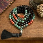 108-bead Buddhist prayer mala bracelet with black onyx, red jasper, and turquoise gemstones, silver Buddha head charm, and black tassel displayed on wooden surface