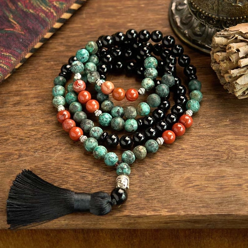 108-bead Buddhist prayer mala bracelet with black onyx, red jasper, and turquoise gemstones, silver Buddha head charm, and black tassel displayed on wooden surface