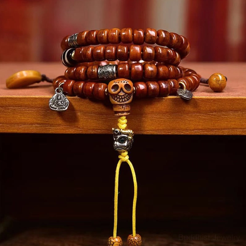 Stacked cylindrical brown bone beads with skull guru bead, silver pendants, yellow tassel displayed on wooden shelf with meditation accessories