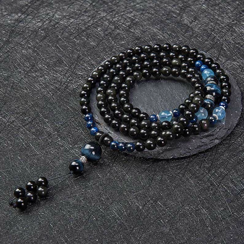 Neatly coiled 108 bead obsidian mala with blue accent stones and triple-bead tassel cluster on dark textured surface