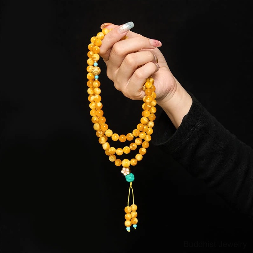 Amber mala prayer beads held in hand wearing black sleeve against black background, showing yellow cord and turquoise tassel