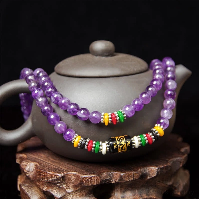 Amethyst mala bracelet with six-word mantra metal beads and colorful accents, side view on wooden carved base with teapot prop