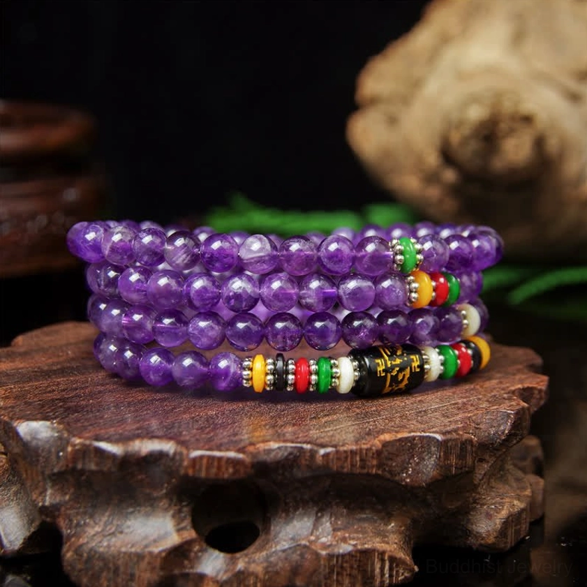 Purple amethyst prayer mala beads with translucent stones and metal mantra decorations displayed on wooden carved base with green plants