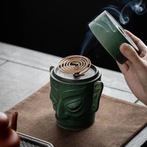 Ancient mask ceramic incense burner with green speckled glaze, overhead view with smoke rising from coil incense, positioned on wooden tea table