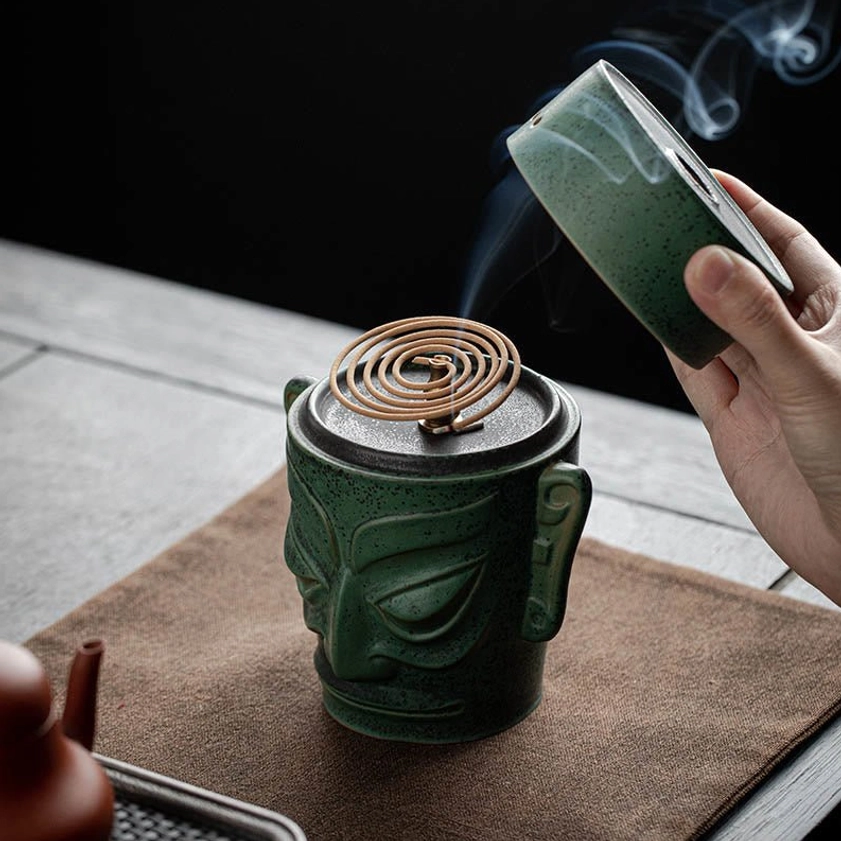 Ancient mask ceramic incense burner with green speckled glaze, overhead view with smoke rising from coil incense, positioned on wooden tea table