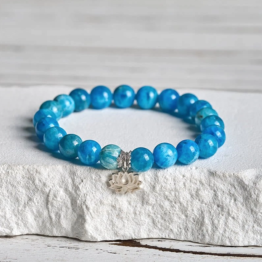 Natural blue apatite lotus sterling silver bracelet displayed on stone surface, showing vibrant round beads with polished silver lotus charm pendant