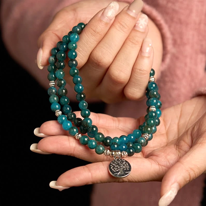 Apatite meditation bracelet two-hand palm display with dark teal beads and silver Tree of Life pendant on dark studio background