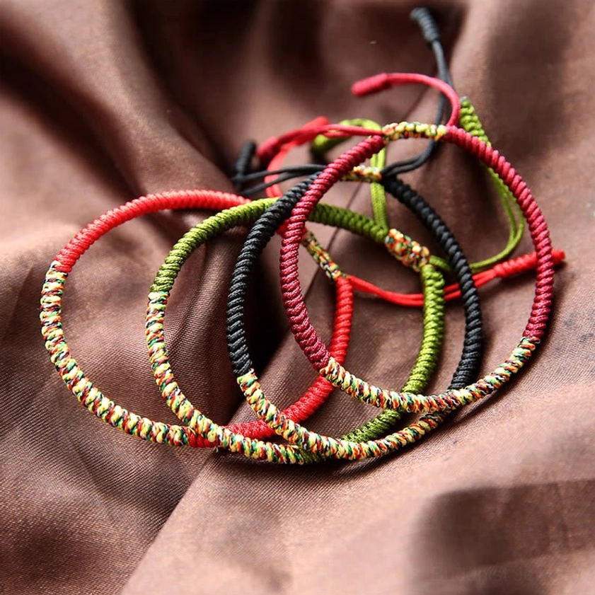Multiple assorted multicolor woven Buddhist bracelets stacked on brown satin fabric, showing various red, black, and green hand-woven designs