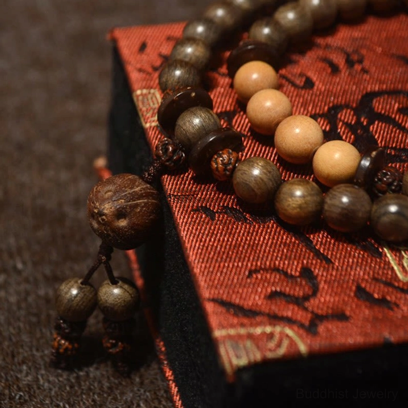 Sacred sandalwood Buddhist mala prayer bracelet with mixed dark and light brown wooden beads and detailed tassel charm, resting on red patterned fabric box
