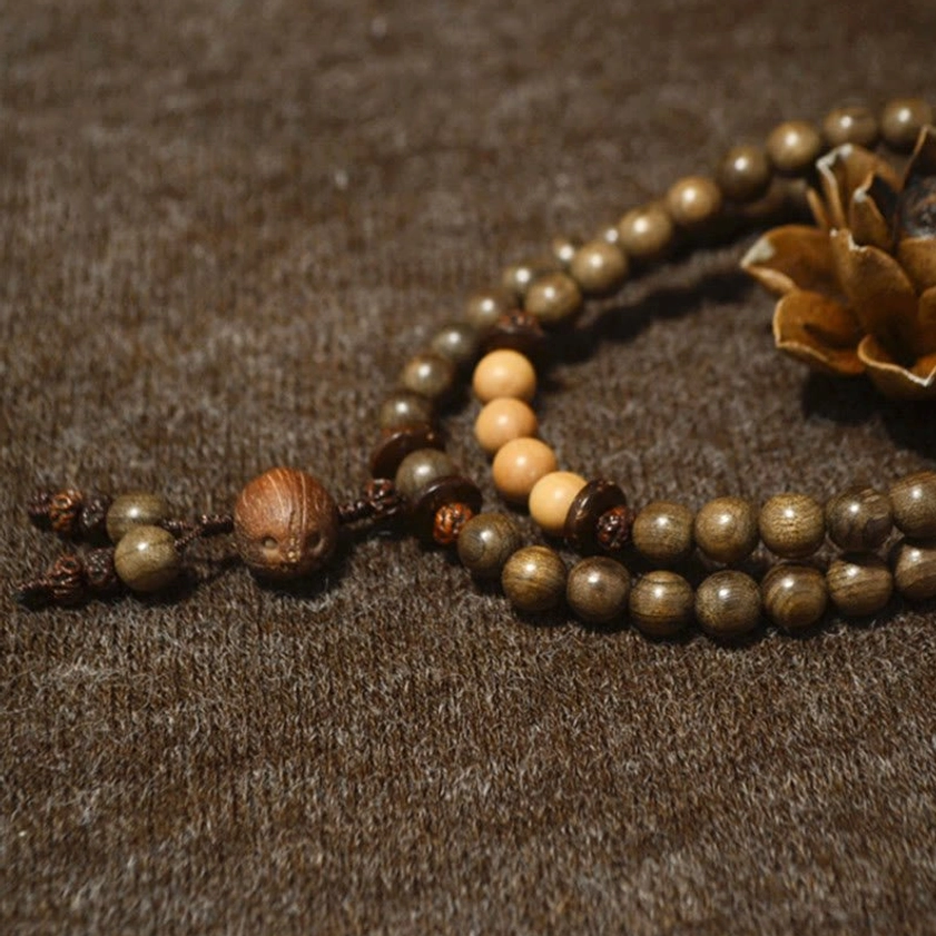 Long sandalwood Buddhist prayer mala with two-tone wood beads and large wooden tassel charm, displayed on brown fabric with dried flowers