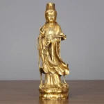 Golden Avalokitesvara brass statue frontal view on wooden desk, showing detailed lotus base and flowing robes with warm polished finish