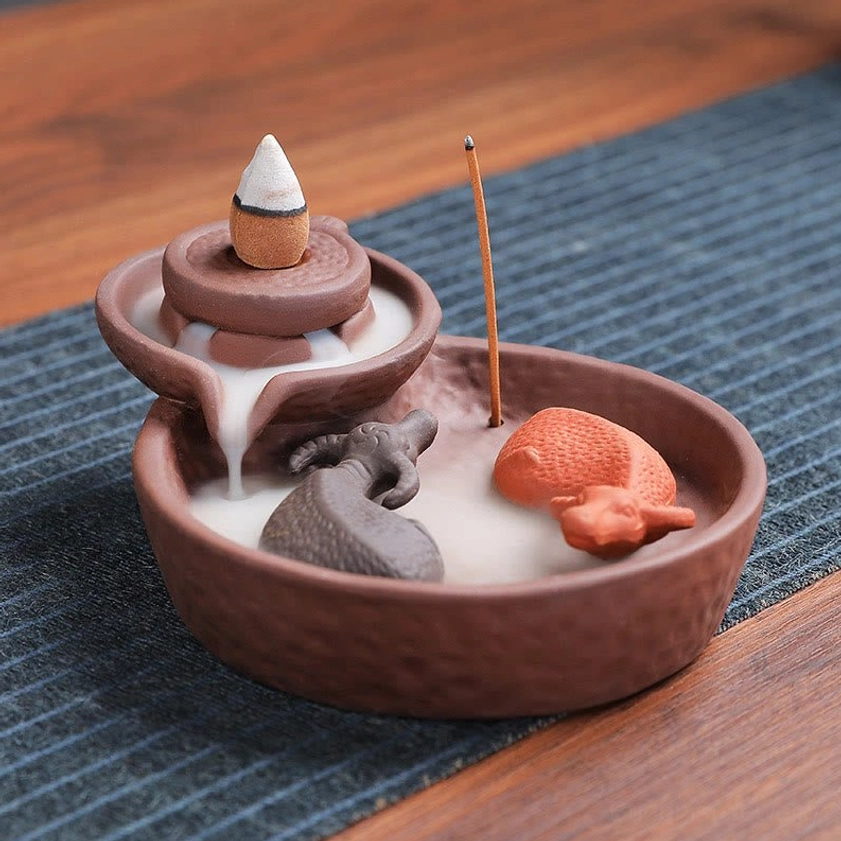 Purple clay backflow incense burner showing flowing smoke cascade effect, mill-shaped holder on wooden table with three-quarter front angle