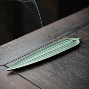Dark green bamboo leaf ceramic incense burner with burning incense and rising smoke on wooden tea table