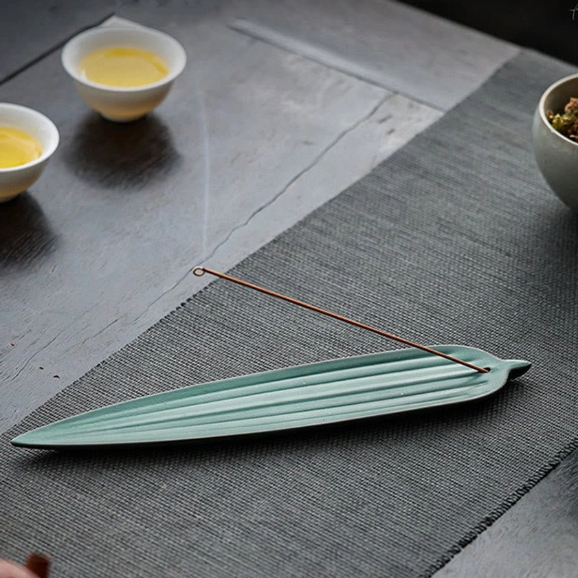 Overhead view of bamboo leaf incense holder with unlit incense stick and tea bowls on woven placemat