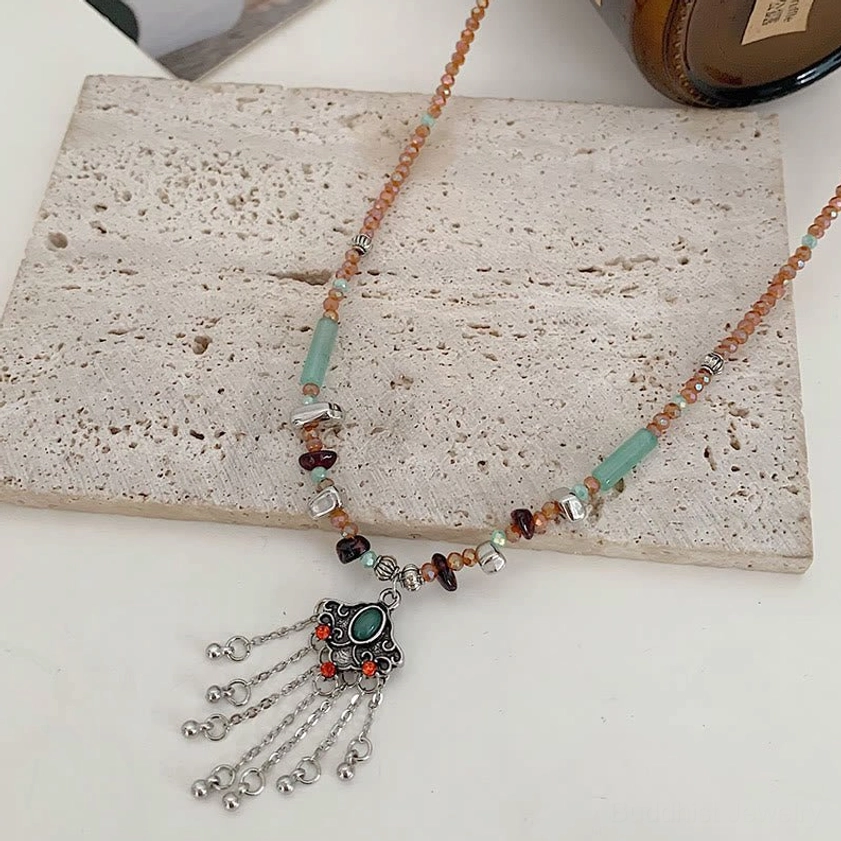 Flat lay top-down view of bohemian beaded tassel necklace displaying brown-red gemstone beads, green stone accents, and silver tassel charm on textured stone surface
