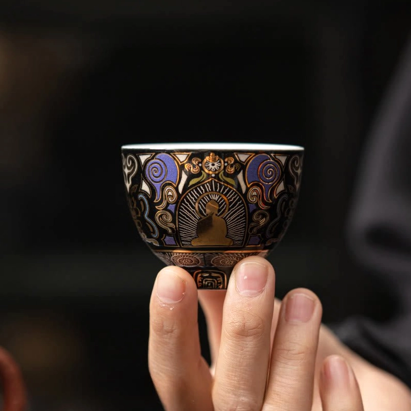 Front view of black ceramic Buddhist tea cup held in human hand showing full decorative Buddha and auspicious cloud patterns