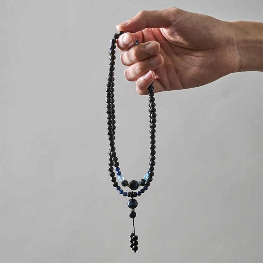 Single strand black obsidian 108 bead Buddhist mala with blue accent beads and hanging tassel pendant on white background