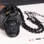 Black obsidian Avalokitesvara Kwan Yin pendant necklace with gold mantra bead displayed on desktop with cast iron teapot, top-down view