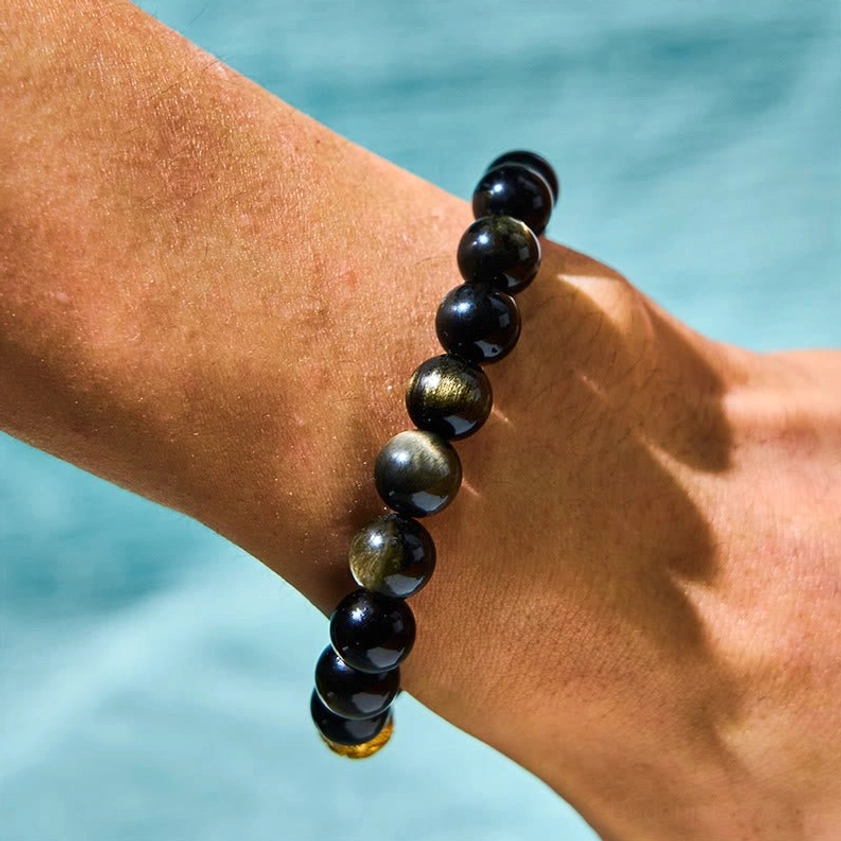 Natural obsidian beaded Buddhist bracelet on wrist, back view showing jet black beads with golden accent details on light blue background