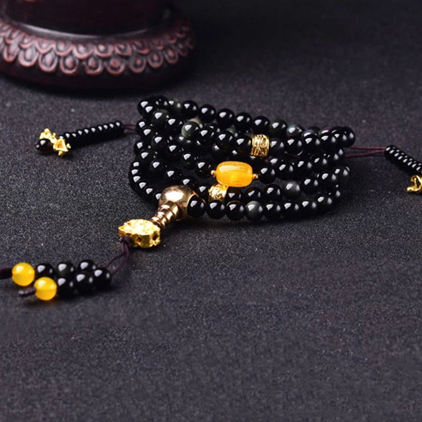 Black obsidian Buddhist mala bracelet laid flat on dark background showing full bead length with gold tassel charms