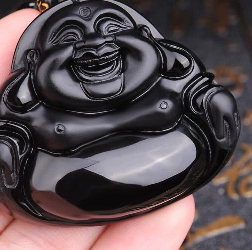 Low-angle close-up of Black Obsidian Buddha pendant held between fingertips, showing detailed facial carving and high-gloss finish