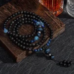 Black obsidian 108 bead Buddhist meditation mala with blue accent stones, displayed on wooden coaster with flat lay styling