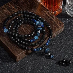 Black obsidian 108 bead Buddhist meditation mala with blue accent stones, displayed on wooden coaster with flat lay styling