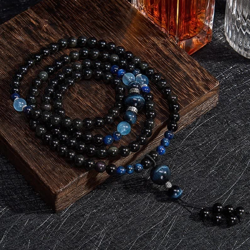 Black obsidian 108 bead Buddhist meditation mala with blue accent stones, displayed on wooden coaster with flat lay styling