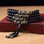 Black Obsidian Om Mani Padme Hum Mala Bracelet with 108 beads, golden engraved script beads, and silver metal accents displayed on red wooden stand