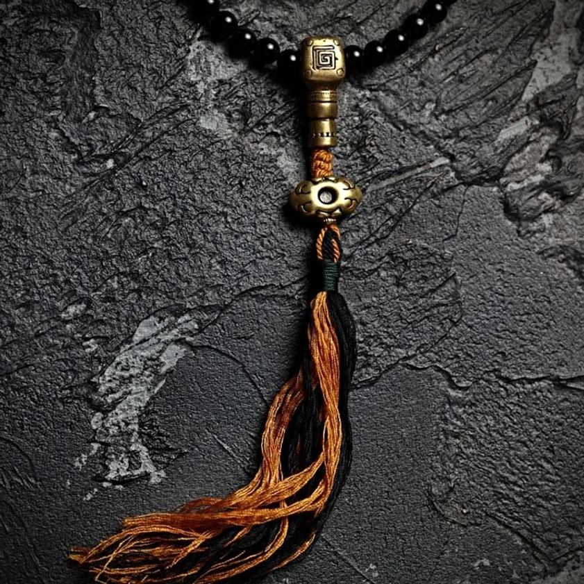 Close-up of black onyx prayer beads with brass guru bead and orange-black tassel on dark stone backdrop