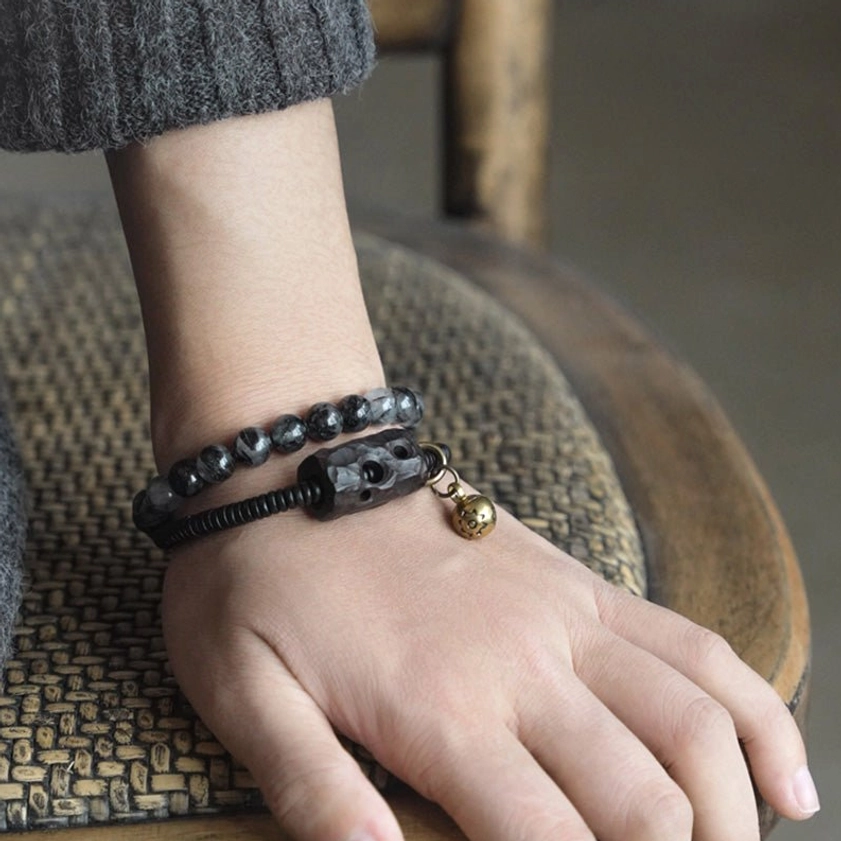 Two matching couple bracelets on hands resting on woven rattan chair, black and smoky gray beads with antique bronze accents