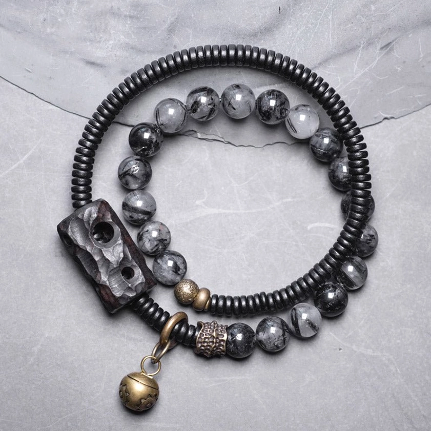 Black rutilated quartz ebony wood copper couple bracelet with carved charm and brass bell pendant on gray surface, top-down view