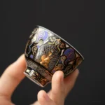 Black Thangka Buddha auspicious clouds ceramic tea cup held in hand, showing intricate gold and purple Buddhist patterns on glossy black ceramic