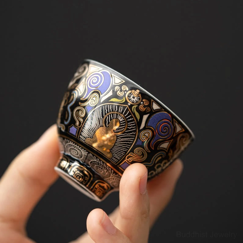 Black Thangka Buddha auspicious clouds ceramic tea cup held in hand, showing intricate gold and purple Buddhist patterns on glossy black ceramic
