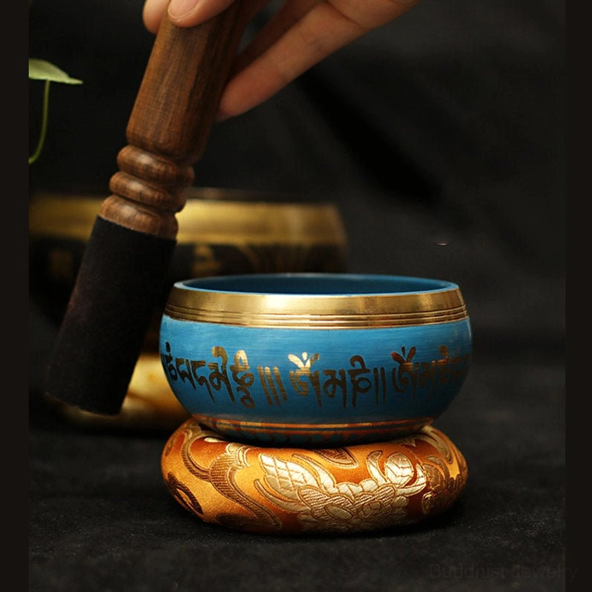 Blue Buddhist singing bowl with gold Tibetan script, held with wooden striker ready to play, positioned on orange embroidered cushion