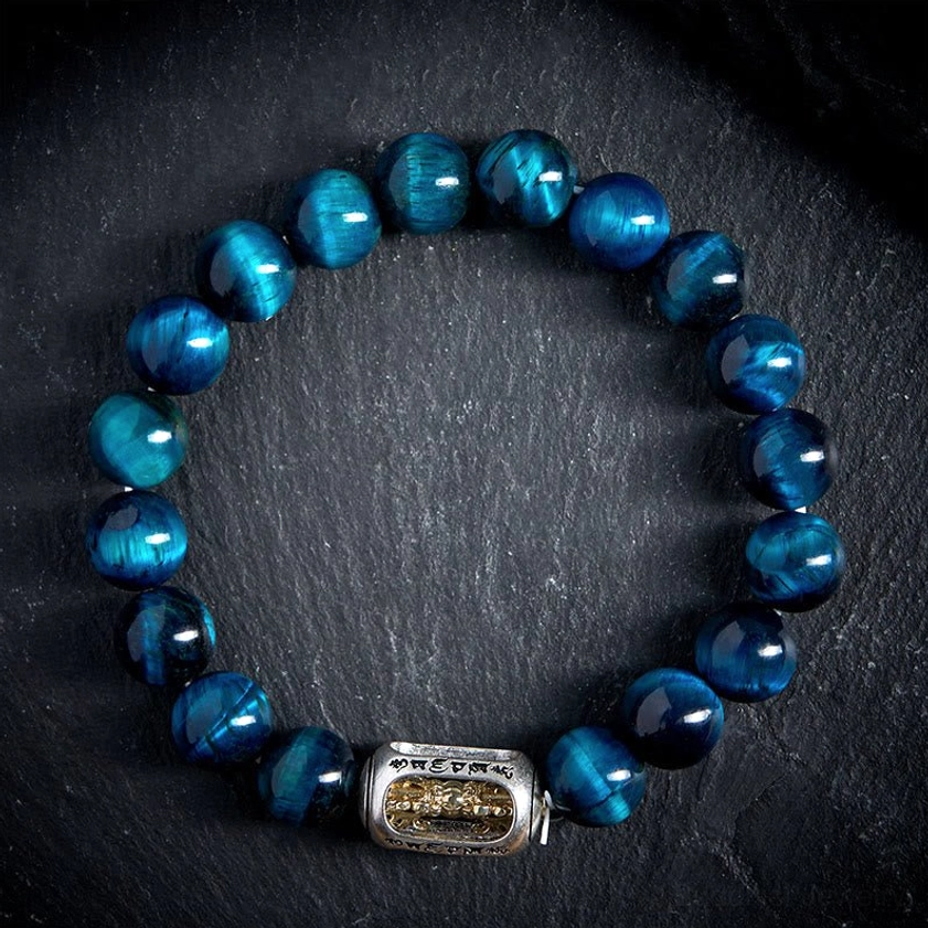 Flat lay top-down view of blue tiger eye Buddhist bracelet showing polished round beads and central engraved mantra charm on black background