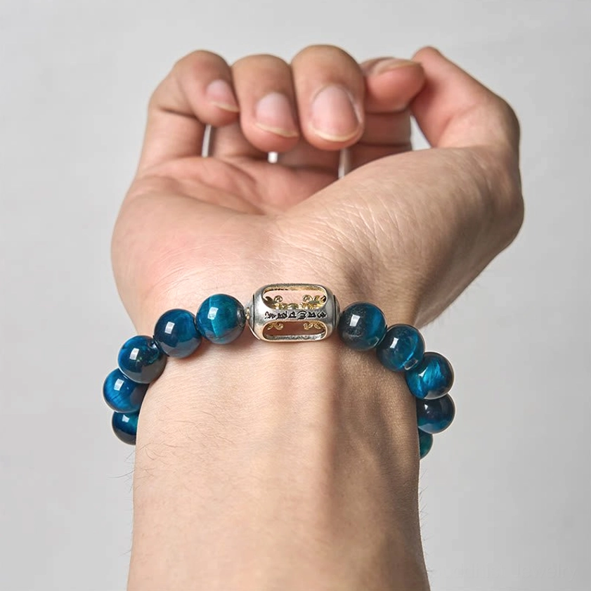 Blue tiger eye beaded bracelet worn on man's hand showing detailed metal engraved Om Mani Padme Hum bead against light grey background