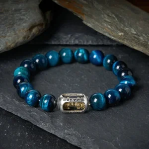 Blue tiger eye Om Mani Padme Hum bracelet with natural cat-eye beads and engraved metal charm displayed on dark slate stones