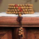 108 bodhi seed mala bracelet with red agate beads stacked display on wooden desk, showing authentic Buddhist prayer beads with spotted brown seeds and braided tassel