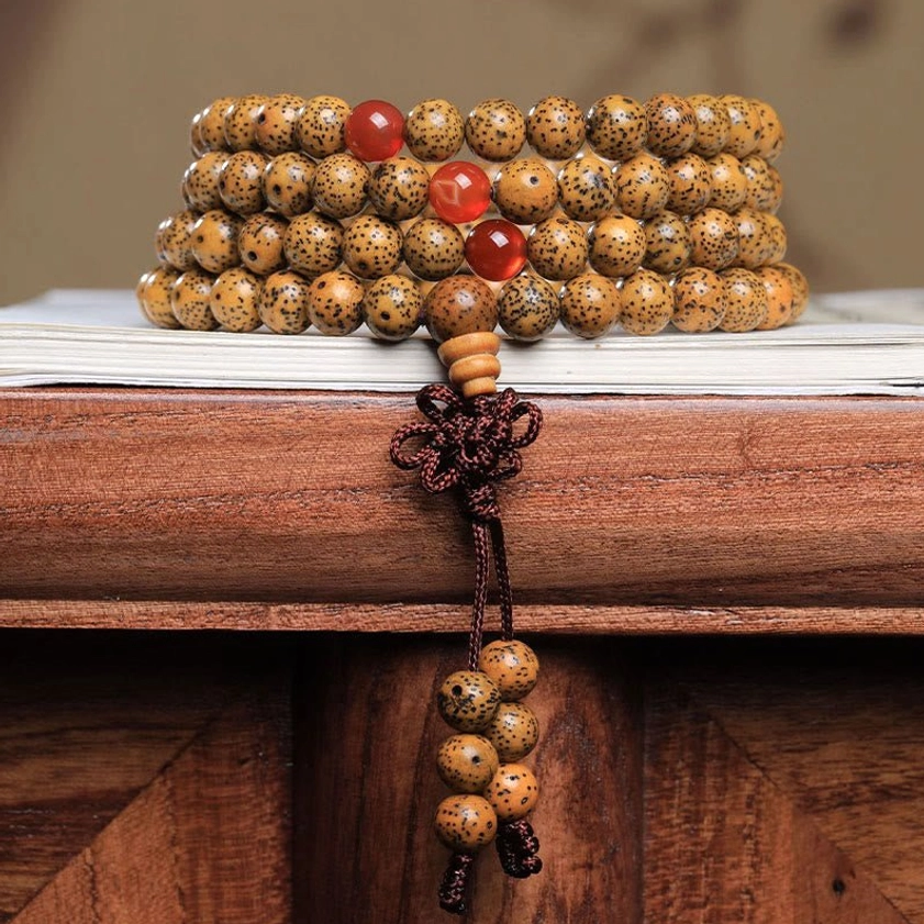 108 bodhi seed mala bracelet with red agate beads stacked display on wooden desk, showing authentic Buddhist prayer beads with spotted brown seeds and braided tassel