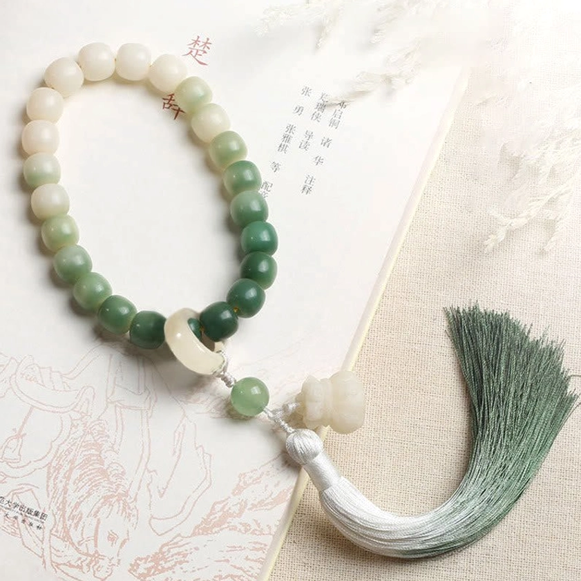 Handmade bodhi seed mala featuring two-tone gradient tassel in dark to light green, ombre beads in white and green tones, and lotus charm on linen cloth backdrop