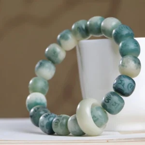 Bodhi seed Om Mani Padme Hum engraved bracelet displayed on white cup and book, showing gradient green-white finish with jade spacer ring