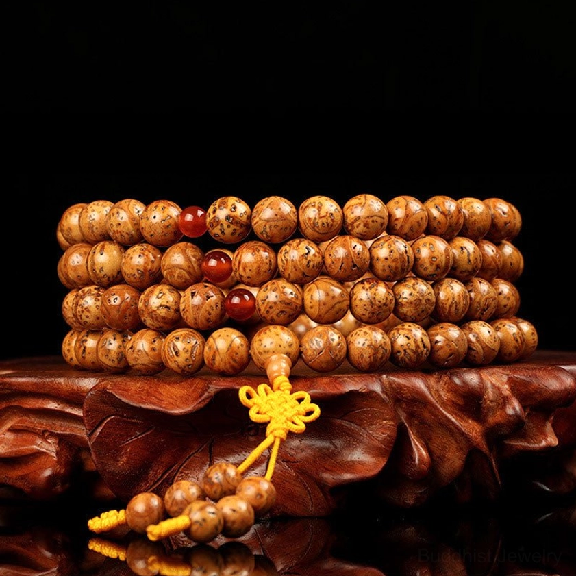 Bodhi seed 108-bead prayer mala bracelet front display on wooden carved base showing tan spotted beads with yellow Chinese knot and red agate spacers