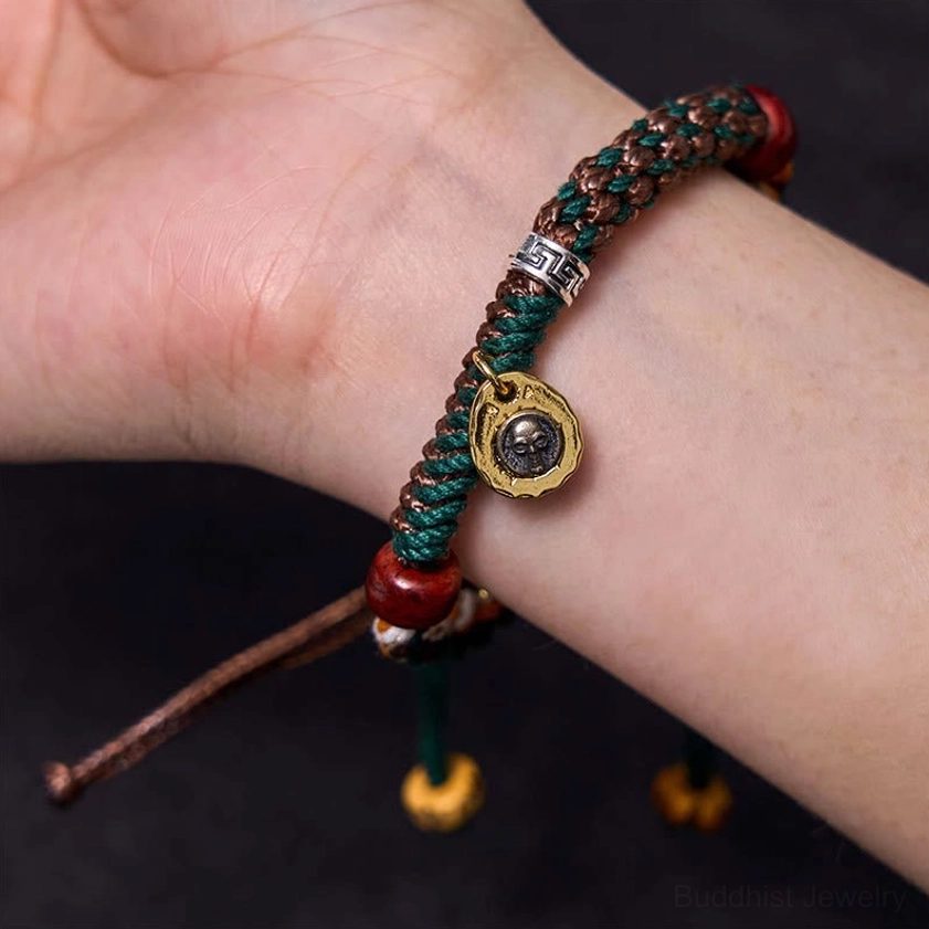 Buddhist bone bead bracelet worn on wrist with green and brown braided rope, red wooden bead, and skull-engraved metal pendant