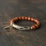 Handcrafted bone beads copper cone pendant strength bracelet displayed overhead on dark rustic metal background, showing brown cylinder beads with antique bronze clasp and spike charm