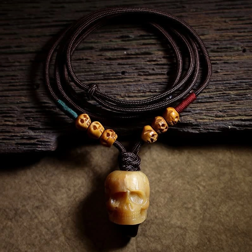 Carved bone skull pendant necklace with braided cord displayed on rustic wooden table, flat lay view