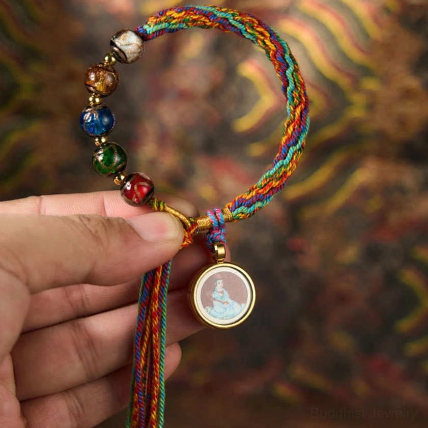 Braided Buddhist blessing string bracelet with multicolor braid, glass beads, round Buddha pendant, and decorative tassel ends on vintage patterned background