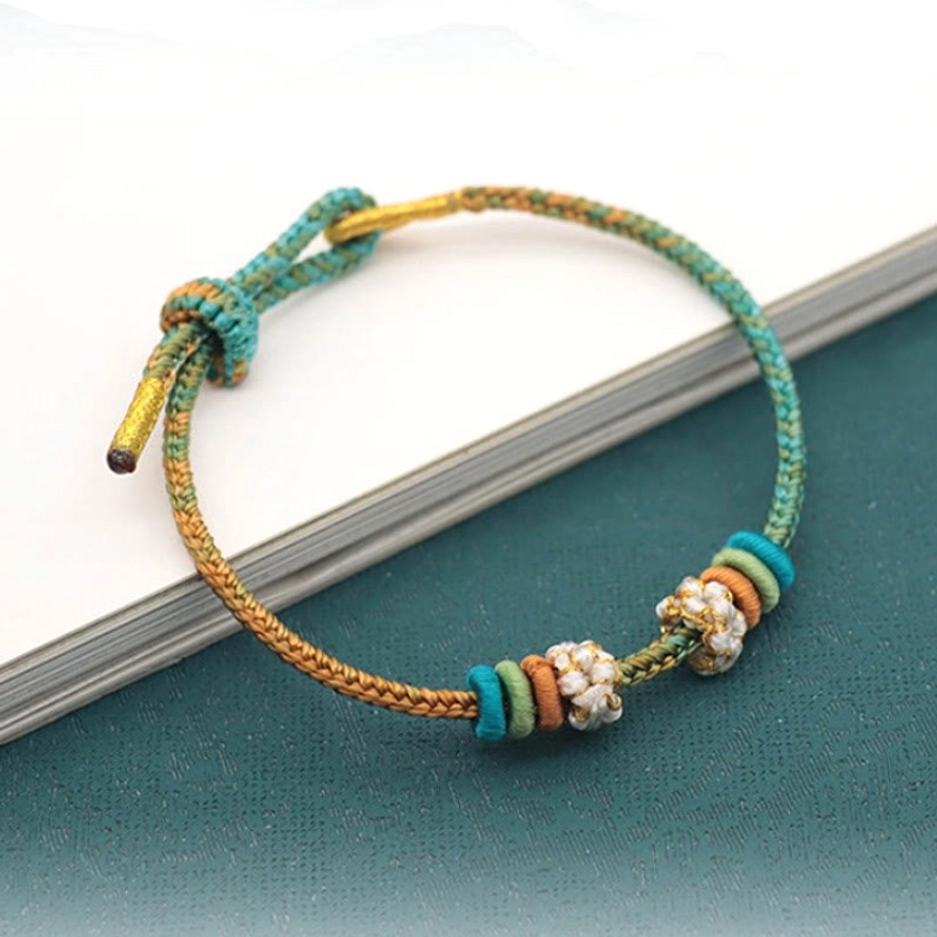 Handmade Buddhist braided bracelet in teal and gold-brown with beaded flower decoration, side view with book backdrop