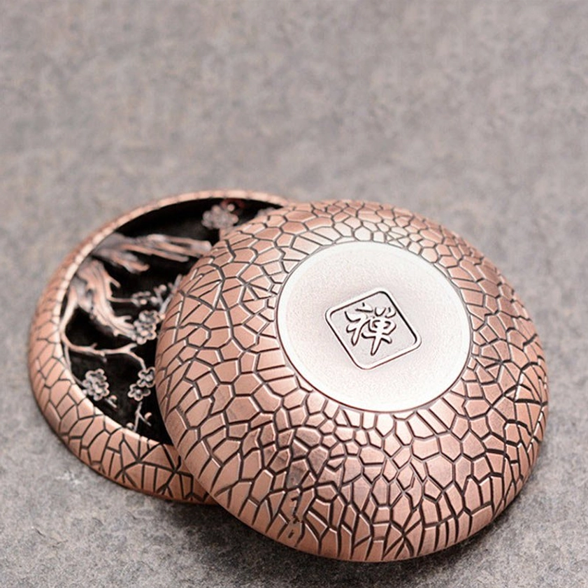 Separated brass incense burner set showing components, zen character engraved on lid back, plum blossom carving on interior, copper red patina on grey fabric surface